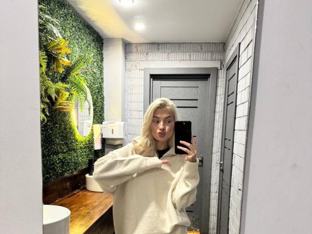 Pearlene Scalley, com cabelo loiro, vestindo um suéter branco e fofo, tirando uma selfie no espelho em um banheiro, segurando um celular preto, com uma expressão séria..