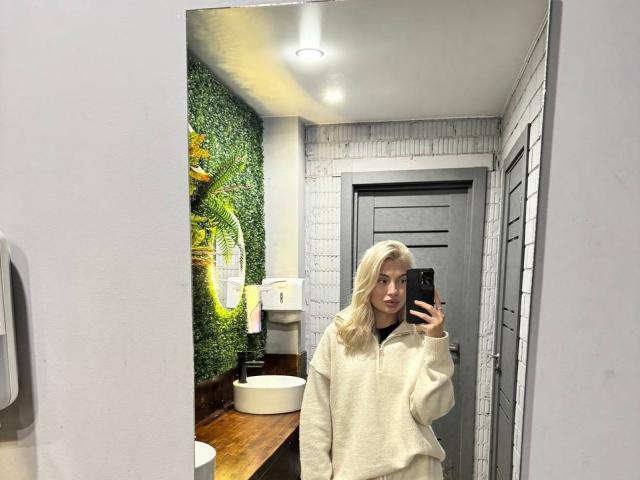 Pearlene Scalley, avec ses cheveux blonds, portant un sweat à capuche blanc, prend un selfie devant un miroir dans une salle de bain, tenant un téléphone noir, debout devant une porte grise..