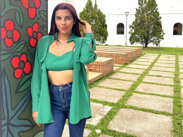 CarlaTorres with green crop top, green shirt, blue jeans, standing by colorful wall, touching hair, medium tits, straight brown hair, white wall, brick pathway, grass, trees.
