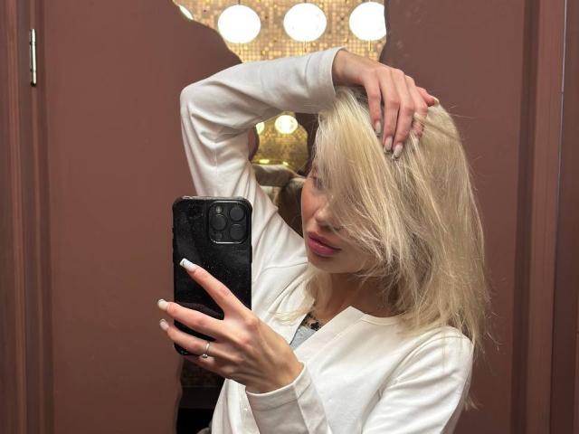 Pearlene Scalley, con i capelli biondi, indossa una camicia bianca a maniche lunghe, tiene in mano un telefono nero, ha una mano tra i capelli, labbra rosa, unghie con le punte bianche, è in piedi di fronte a uno specchio, con pareti marroni..