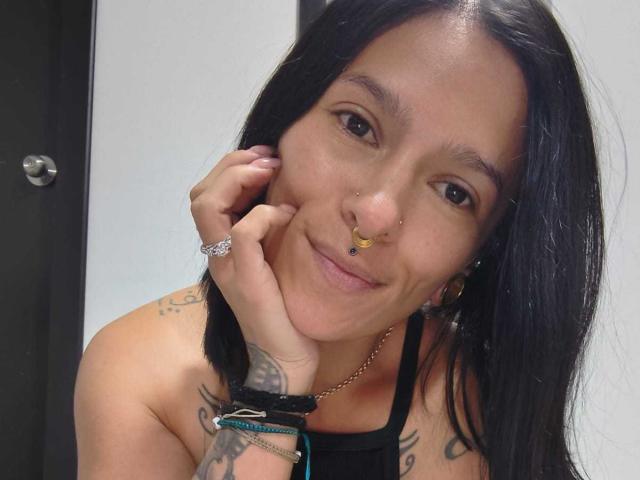 AmelieBlake with black hair, nose ring, and tattoos, smiling at the camera, hand on cheek, wearing a black halter top, earrings, and bracelets.