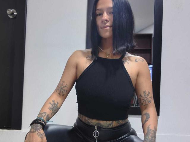 AmelieBlake with black bob, nose ring, black halter top, tattoos on arms and belly, black leather pants, sitting, confident, slight smile, wearing black wristbands.