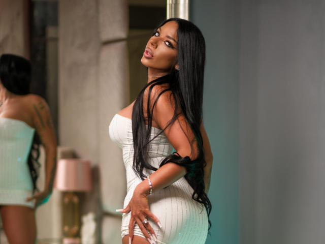 SonyaStorm, com longos cabelos pretos, vestindo um vestido branco justo, exibindo seu grande traseiro e pequenos seios, olhando para trás com uma expressão emburrada, com as mãos no bumbum..