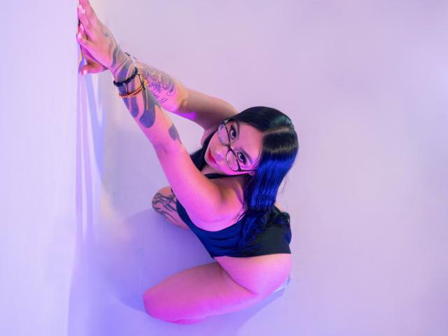 ChloeMoretti inginocchiata, in un costume intero nero, braccia tatuate, occhiali, capelli blu-neri, sorridente, mani appoggiate su un muro bianco, illuminazione viola-rosa, che guarda verso l'alto, mostrando il suo sedere..