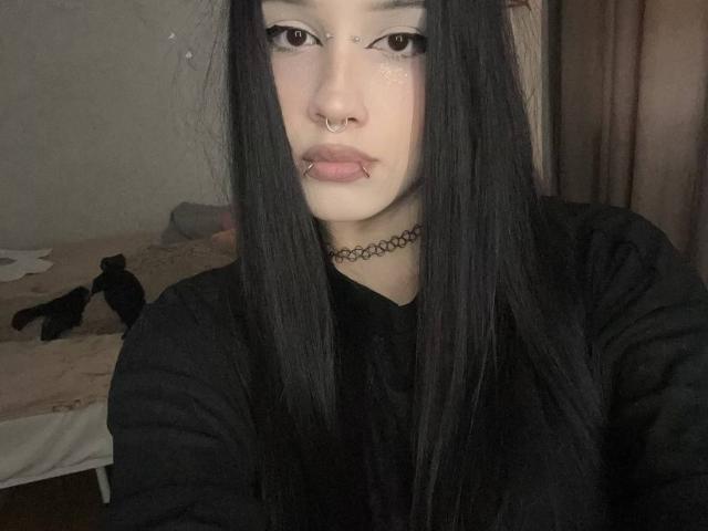 LadyKezele, com cabelos pretos longos, pele clara, piercing no nariz, piercing no lábio, colar preto e camisa preta, olhando para a câmera, em um quarto com pouca luz..