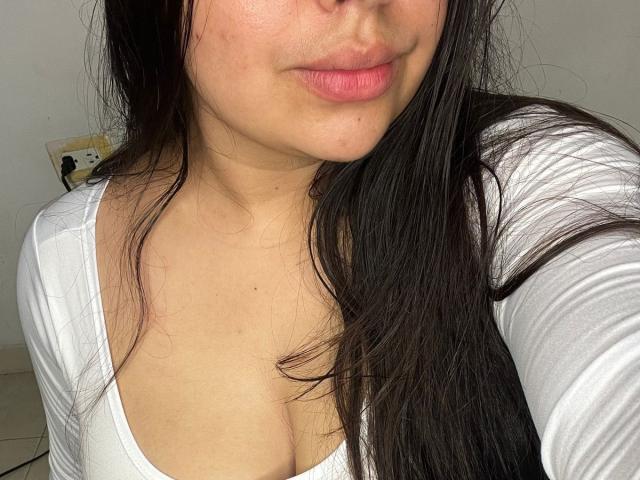 SammyTitss with long dark hair, pink lips, and light skin, wearing a white top with a low neckline, showing her small tits and slightly visible cleavage in a selfie.