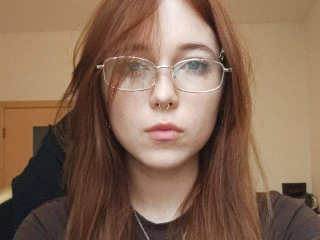 ArlenaStinnett with red hair, glasses, nose ring, and pouty lips, wearing a black shirt, looking straight at the camera. Her blue eyes are slightly squinted.