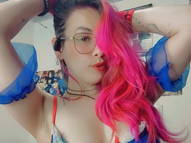 NatashaLau, avec ses cheveux roses, ses lunettes et son rouge à lèvres rouge, les bras levés, portant des manches transparentes bleues, des bretelles de soutien-gorge rouges et un collier, montrant ses seins et ses tatouages..