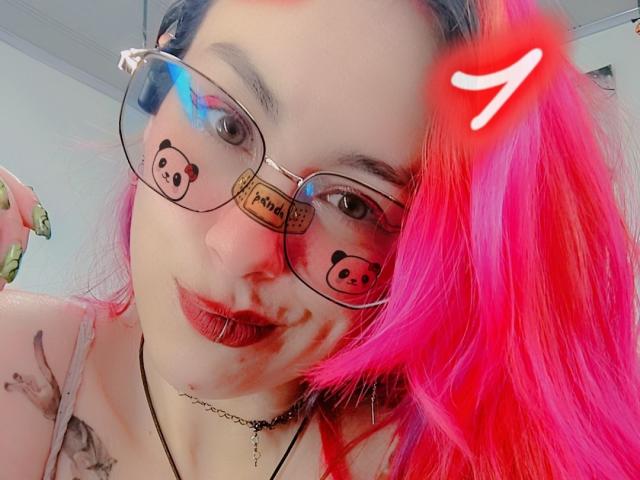 NatashaLau, avec ses cheveux roses, portant des lunettes avec des motifs d'ours, du rouge à lèvres rouge et un collier noir, a un tatouage sur son épaule, et le chiffre 7 en haut à droite..