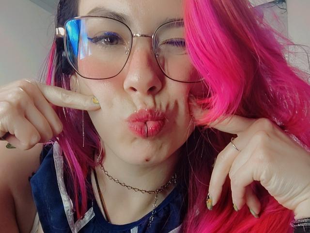 NatashaLau, avec ses cheveux roses et noirs, ses lunettes et ses lèvres pulpeuses, pointe du doigt près de son visage, portant une chemise bleue, une bague et un collier, et faisant une grimace dans un autoportrait..