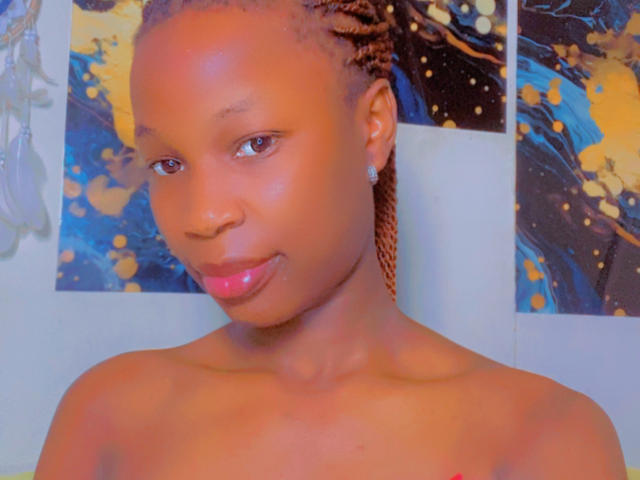 JazzBerry tiene una piel marrón lisa, pequeños aretes redondos, y sus trenzas están recogidas. Está sin ropa, con lápiz labial rosa y una sonrisa sutil. Sus ojos marrones miran hacia adelante..