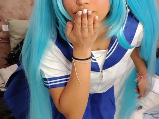 YayaLm with long blue hair, white and blue schoolgirl outfit, hand covering mouth, white nails, ring on finger, black bracelet, pink necklace, leaning forward, bedroom setting.