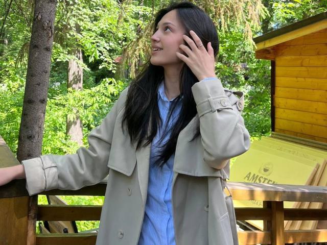 AdrianneRossey with long black hair, wearing a beige coat over a blue shirt, leaning on a wooden railing, talking on a phone, with green forest background.