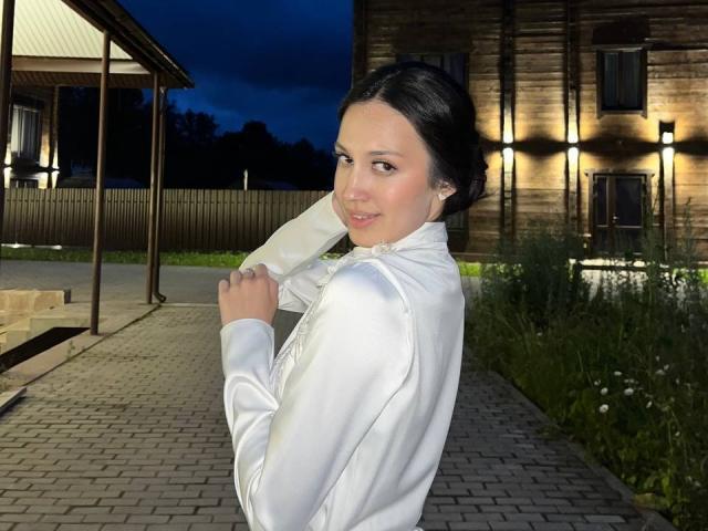 AdrianneRossey with dark hair, white shirt, and subtle smile, looking over shoulder, holding shirt, showing her small, perky tits, in a nighttime setting with a wooden building.
