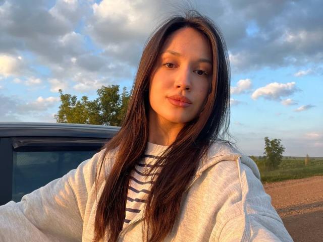 Adrianne Rossey, met lang bruin haar, die een witte hoodie en een gestreepte shirt draagt, heeft een schattig gezicht en een lichte glimlach, en staat naast een auto, met een zonnige, bewolkte lucht op de achtergrond..