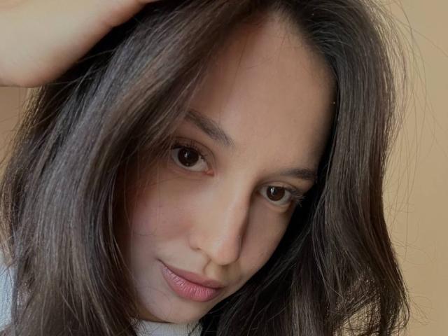 AdrianneRossey has wavy brown hair, brown eyes, and a slight smile with pink lips. She's touching her hair with one hand, showing a cute and relaxed expression.