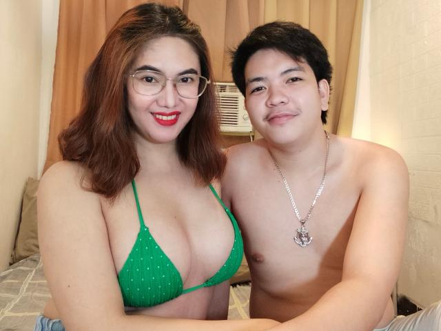 AsianCoupleDesire con grandi seni, in un bikini verde a pois, e AsianCoupleDesire con una collana d'argento, entrambe sorridenti, sedute su un letto. AsianCoupleDesire ha i capelli neri corti..