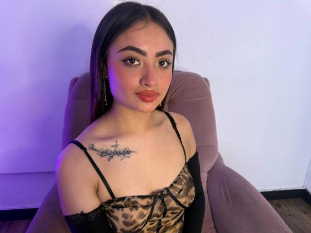 Samantha Coppers, met lang donker haar, draagt een top met luipaardprint en zwarte bandjes, heeft een tatoeage op haar rechterborst en zit in een roze stoel, met een glimlach..