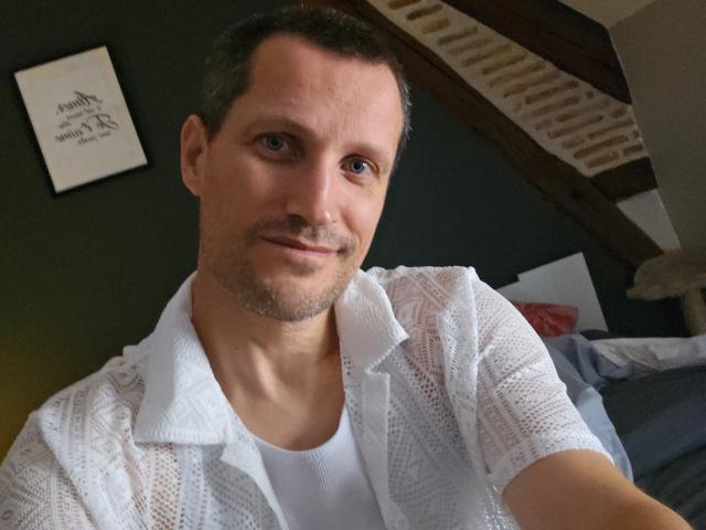 MisterFredX, com cabelo castanho curto, barba rala e uma camisa de malha branca por cima de uma camiseta branca, sorrindo levemente. Seus olhos azuis estão fixos na câmera..