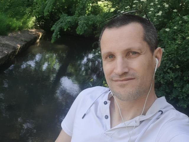 MisterFredX, com cabelo castanho curto, camisa branca e fones de ouvido, sorrindo perto de um riacho escuro e reflexivo, cercado por folhagem verde..