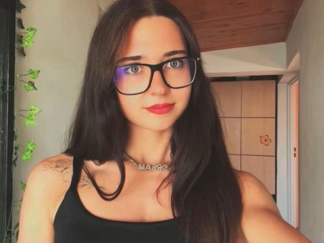 VanessaShineX with long dark hair, black glasses, red lipstick, black tank top, silver necklace, and visible tattoo on her right shoulder, smiling at the camera.