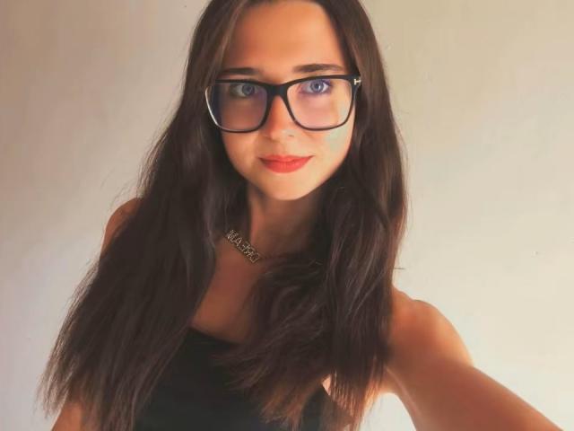 VanessaShineX with long dark brown hair, black-framed glasses, blue eyes, red lipstick, and black sleeveless top, showing her tits slightly. She's smiling at the camera.