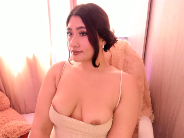 ElyRoss with dark hair in a bun, wearing a beige, strapless top, showing her big, round boobs, sitting in a pink, fluffy chair, smiling softly.