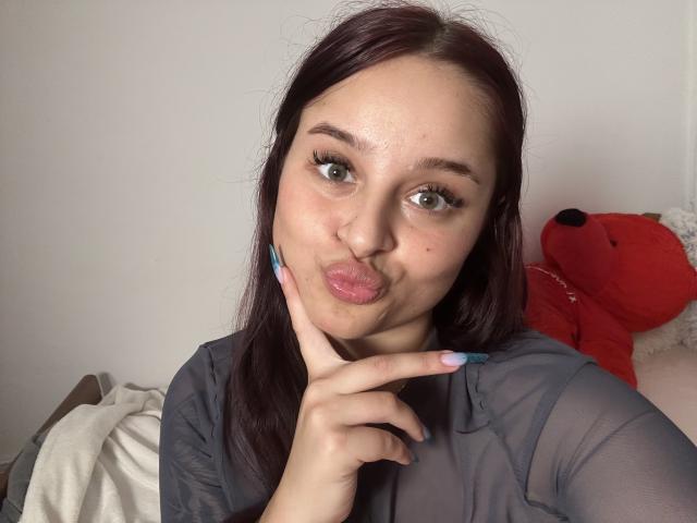 Lucie: Roodharig, met volle, roze lippen, groene ogen en donkerbruin haar, gekleed in een doorschijnend grijze top, die haar kin aanraakt met een vinger, naast een rode knuffelbeer..