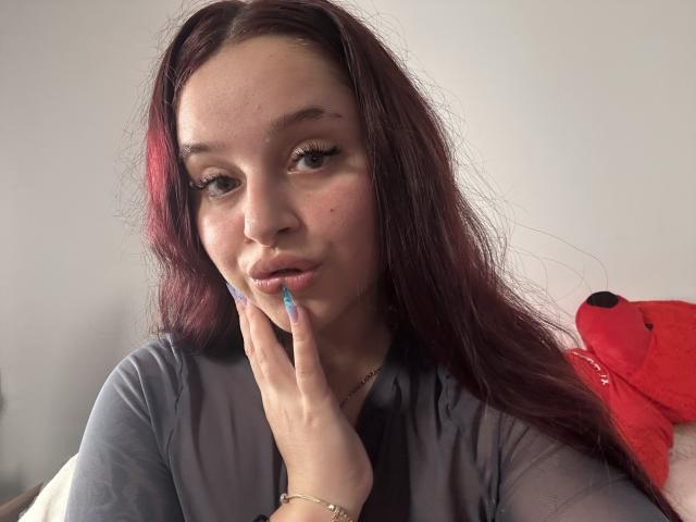 Lucie: Rotes Haar, das lang gefärbt ist, nachdenklicher Gesichtsausdruck, helle Haut, trägt ein graues Hemd, berührt ihr Kinn mit blau lackierten Nägeln, leichte Röte, goldenes Armband und ein rotes Stofftier..