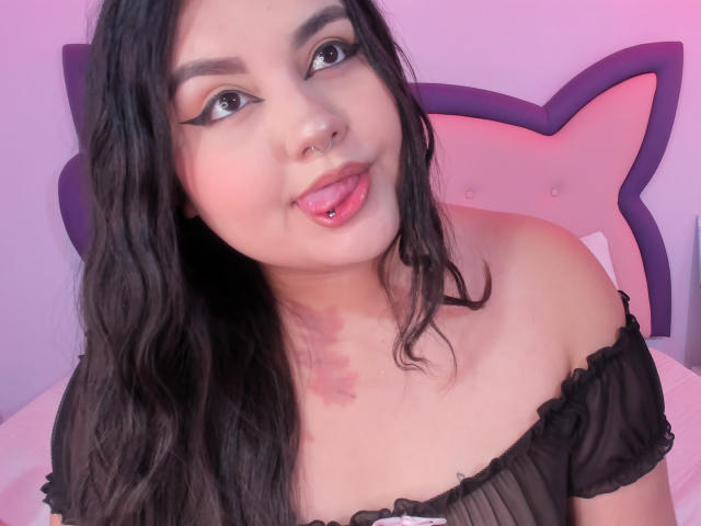 MarlaBriard with black wavy hair, nose ring, and pink tongue piercing, wearing an off-shoulder black top, looking up with makeup and a slight smile.