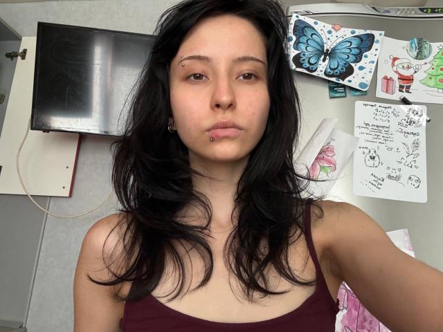 Desiree Rampulla a les cheveux noirs, un piercing au nez et un piercing aux lèvres, et elle porte un débardeur bordeaux qui révèle ses seins nus, avec une expression neutre et légèrement boudeuse..