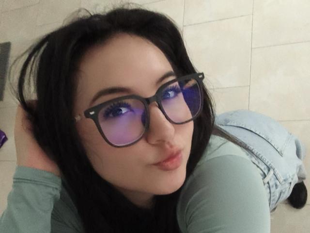 Susan Monroy mit dunklem Haar, blauen Gläsern, die Licht reflektieren, voller Lippen, einem grünen Hemd und hellblauen Jeansoveralls, liegt auf einem gefliesten Boden, die Hand im Haar, und schaut in die Kamera..