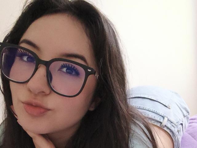 Susan Monroy, con lunghi capelli scuri, indossa occhiali neri, guarda la fotocamera con un riflesso viola, ha occhi azzurri, labbra rosa e pelle chiara, è appoggiata a un letto viola e indossa pantaloncini di jeans chiari..