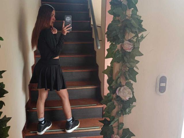 ValerieWoods with long brown hair, black long-sleeve top, black pleated skirt, black-and-white sneakers, taking a selfie on wooden stairs, surrounded by green plant vines.