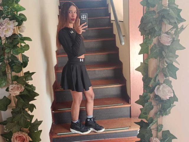 ValerieWoods, avec de longs cheveux bruns, un haut et une jupe noirs Under Armour, des baskets noires, debout sur des marches en bois, prenant un selfie devant un miroir, montrant ses fesses, avec une décoration de feuilles vertes et de roses..