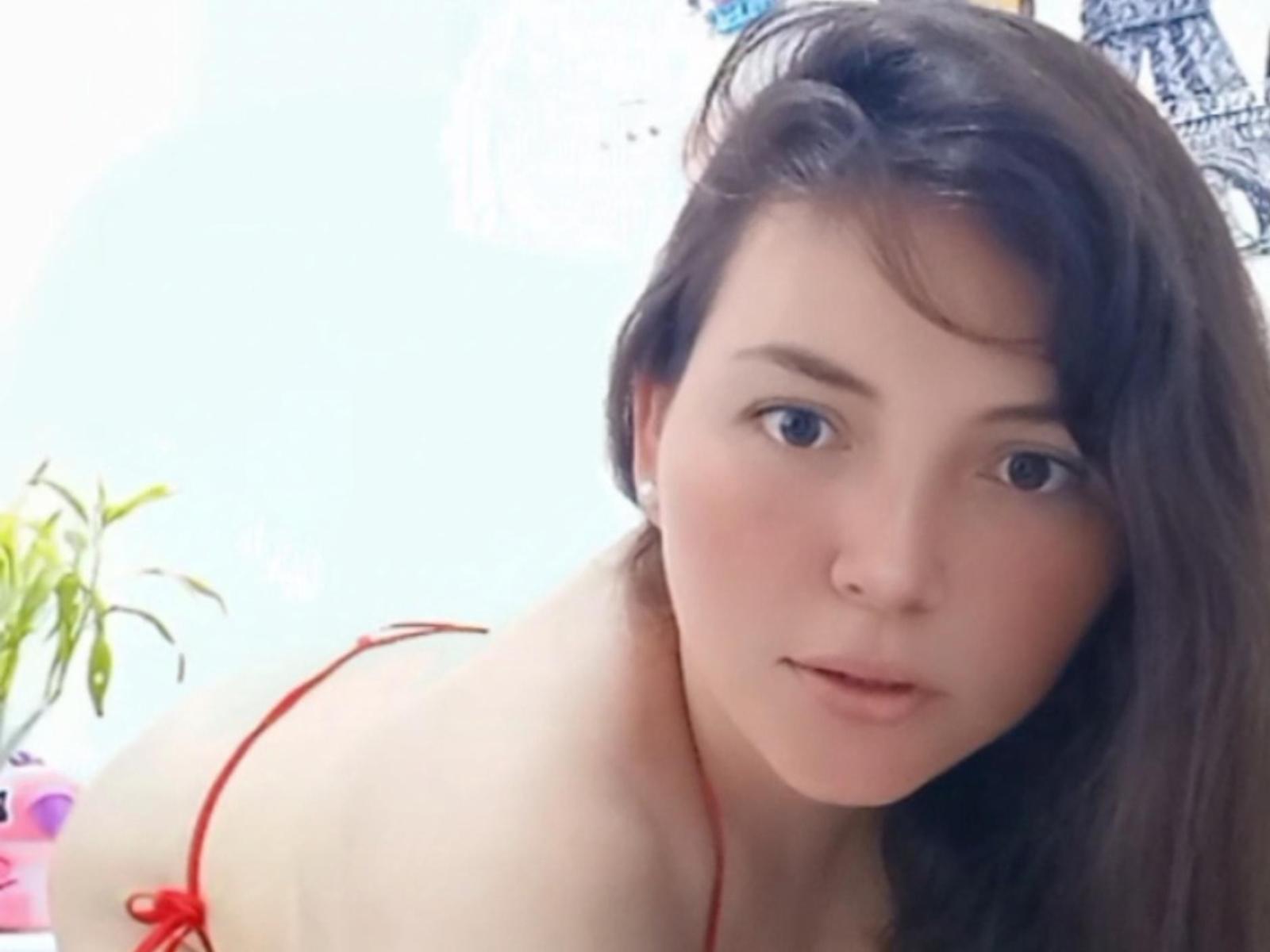 Kahtya, avec ses cheveux bruns, son bikini rouge, sa peau pâle et ses petites seins, est penchée en avant, regardant l'appareil photo avec un sourire discret. Son vagin est à peine visible..