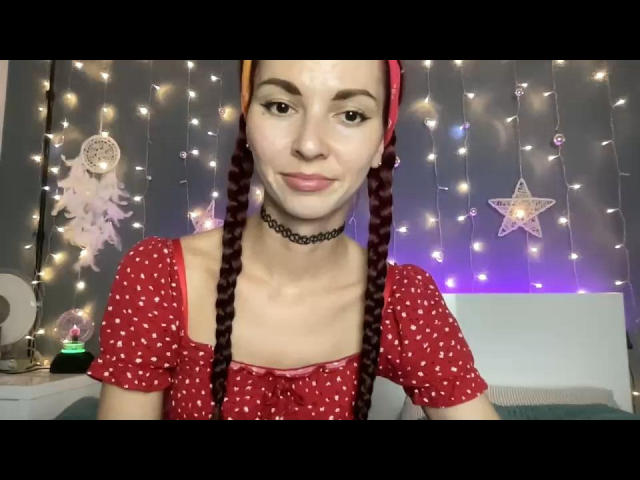 Deine Monica mit geflochtenem, rotem Haar, die ein rotes Kleid mit Punkten und eine schwarze Halskette trägt, hat ein süßes Lächeln, kleine Brüste und eine schlanke Figur, und befindet sich in einem gemütlichen, beleuchteten Raum..