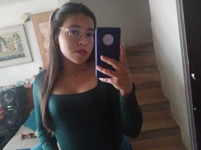 DayannaMora with glasses, long brown hair, green top, holding purple phone, taking mirror selfie, showing her tits, standing on wooden stairs, red lipstick.