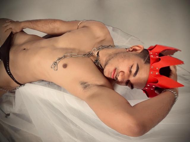 MaikolTwinkX, sin camisa, vistiendo una tanga negra y una corona roja con púas, collar de cadena, acostado sobre tela blanca, con los brazos por encima de la cabeza, mostrando vello axilar, cabello castaño corto, atractivo, piel clara..