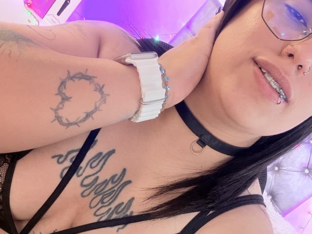 Natalie Harris, com cabelo preto, óculos e piercing no nariz, usando um colar preto e um relógio, mostrando tatuagens no braço e no peito, e seus seios. Luzes rosa e roxa..