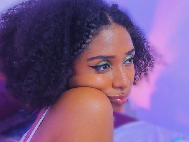 MeganWlliams with dark curly hair, green eyeshadow, and a nose ring, resting her chin on her shoulder, showing off her shiny brown skin and glossy lips.