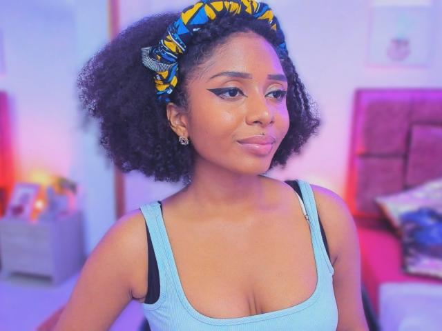 MeganWlliams with curly black hair, wearing a blue and yellow patterned headscarf, light blue tank top, black bra, and eyeliner, smiling, showing her nose ring and small tits.