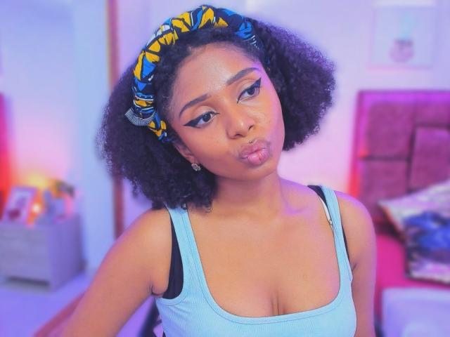 MeganWlliams with curly black hair, wearing a blue headband with yellow patterns, pouting her lips, in a light blue tank top, showing her small tits and black bra.