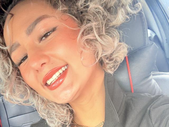 ElenaSaenz with curly grayish hair, red lipstick, and a shiny smile, wearing a black jacket, sitting in a car, head tilted back, eyes half-closed, looking sexy.
