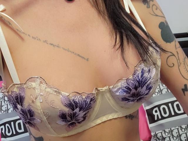 Französisch: Shannas Brüste in einem durchsichtigen, beige farbenen BH mit violetten Blumenstickereien. Sie hat Tattoos auf ihrer Brust und ihren Armen, und ihr schwarzes Haar ist teilweise sichtbar..