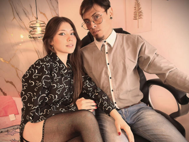 Aeron69, avec de longs cheveux bruns, un t-shirt noir à motifs tourbillons, des bas résille, assise sur une chaise, à côté d'Aeron69 portant des lunettes, un t-shirt gris, un jean bleu, regardant la caméra, dans un bureau..