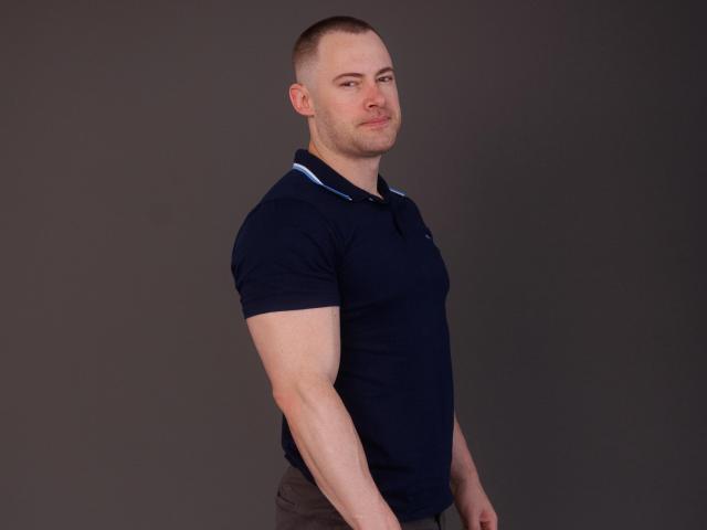 Mhagoo, een gespierde man met een kaalgeschoren hoofd, gekleed in een marineblauwe polo, staat met zijn zij aan de camera en heeft een zelfverzekerde grijns, terwijl hij zijn biceps laat zien..