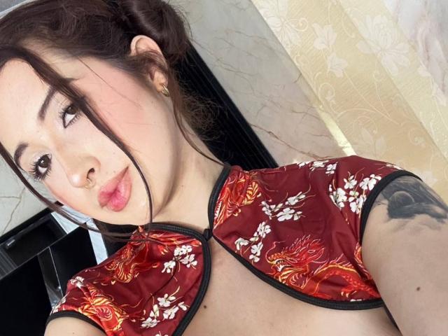 MiaVolkova with Asian features, brown hair in a bun, wearing a red floral Chinese dress, black trim, small tattoo on right arm, pouty pink lips, looking at camera.