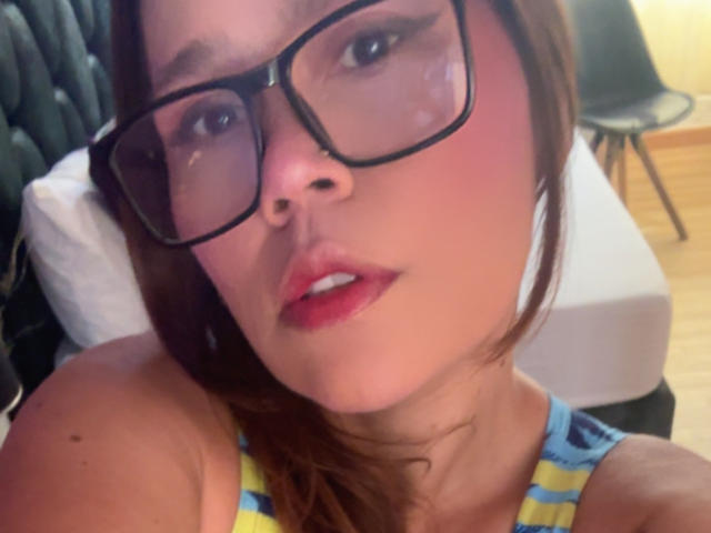 SophiaQueens con gafas, labios rosados y pintalabios rojo, vistiendo una blusa a rayas amarilla y azul, mirando a la cámara con los labios rojos ligeramente entreabiertos..