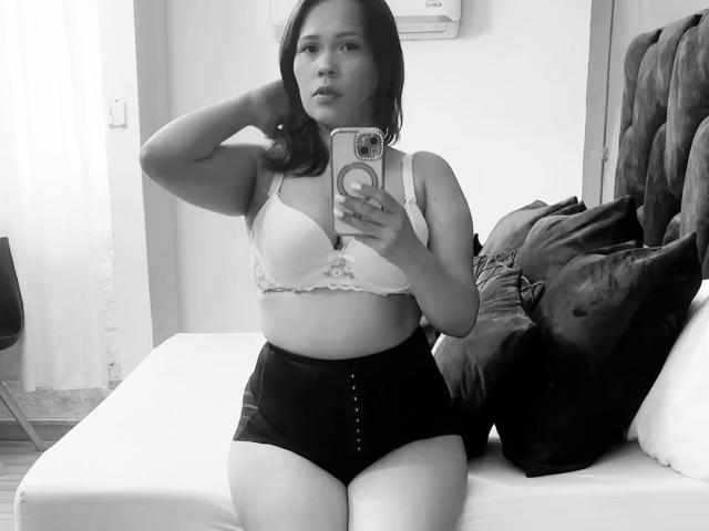SophiaQueens, de figura voluptuosa, con pechos pequeños en un sostén de encaje blanco y tangas negras de cintura alta, tomando una foto de sí misma en el espejo, con el brazo derecho levantado, sosteniendo un teléfono, sentada en una cama..